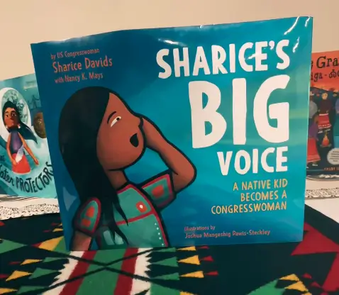 A children's book titled "Sharice's Big Voice: A Native Kid Becomes a Congresswoman" is displayed, featuring an illustrated character on the front cover. Other books and a colorful fabric are in the background.